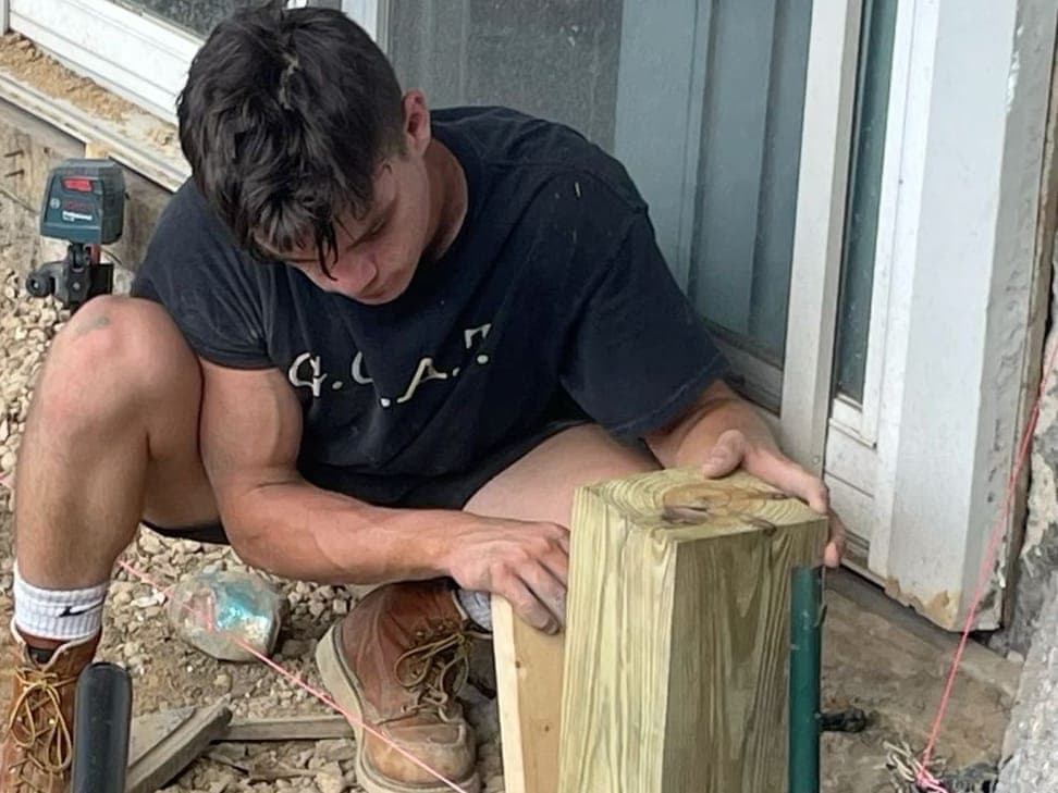 Dominic Dawson in a work shirt and boots sets a pressure-treated post in a gravel trench beside a house window, a string line across the trench.