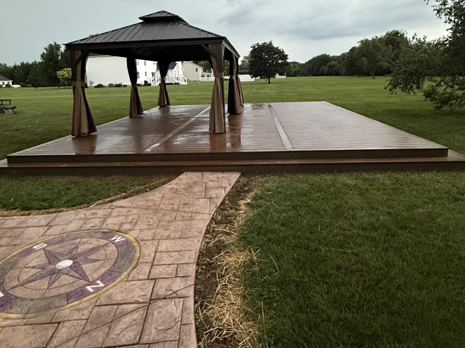 A composite deck with a dark metal gazebo on it and a stamped concrete walkway with a compass rose, in a wide lawn under an overcast sky.